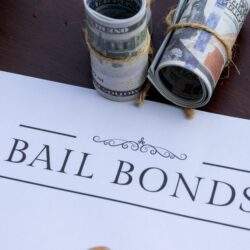 What is Bail - Picture of a piece of paper titled Bail Bonds sitting on a desk with rolls of $100 bills above it & a pen on top of the paper