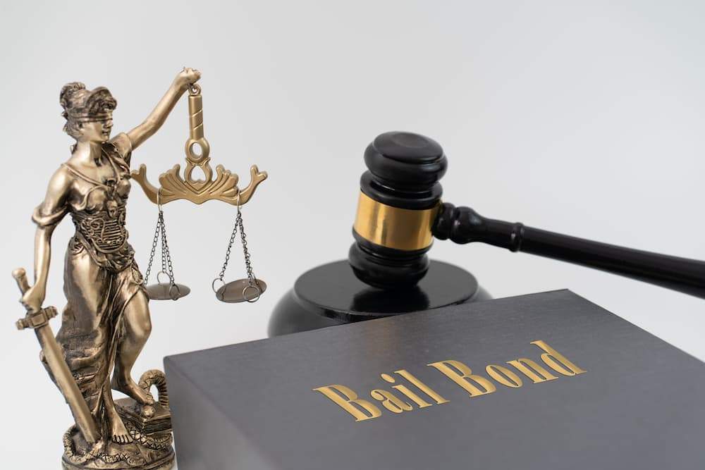 Lady Justice statue, a judge's gavel, and a book labeled "Bail Bond," symbolizing the legal paperwork and processing required to secure and maintain various types of bail bonds.